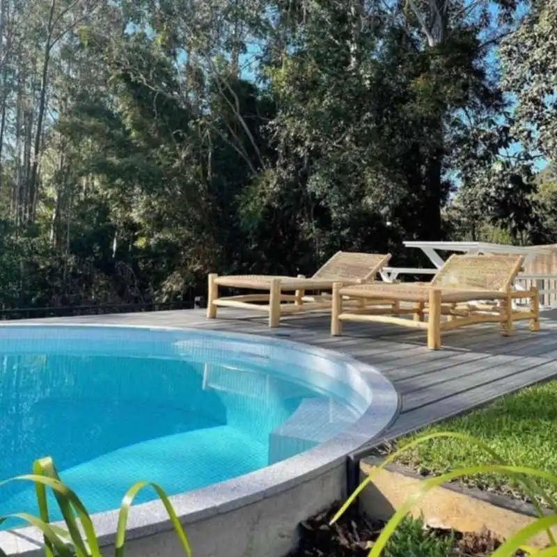 A circular above-ground plunge pool with light blue tiles is built into a grey timber deck. Two woven bamboo sun loungers sit on the deck, overlooking a dense backdrop of tall eucalyptus trees under a clear sky.