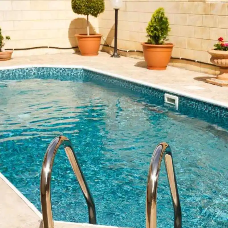 A custom concrete plunge pool features shimmering teal mosaic tiles and a stainless steel ladder. The pool is surrounded by light stone paving and several terracotta pots containing small, neatly trimmed shrubs.
