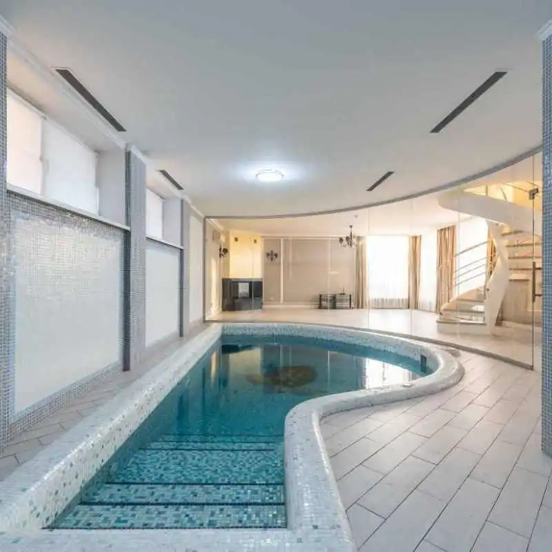 An indoor freeform pool with a curved edge and white mosaic tiling is set within a modern, bright room. The space features white floor tiles, a glass partition wall, and a white spiral staircase in the background leading to an upper level.