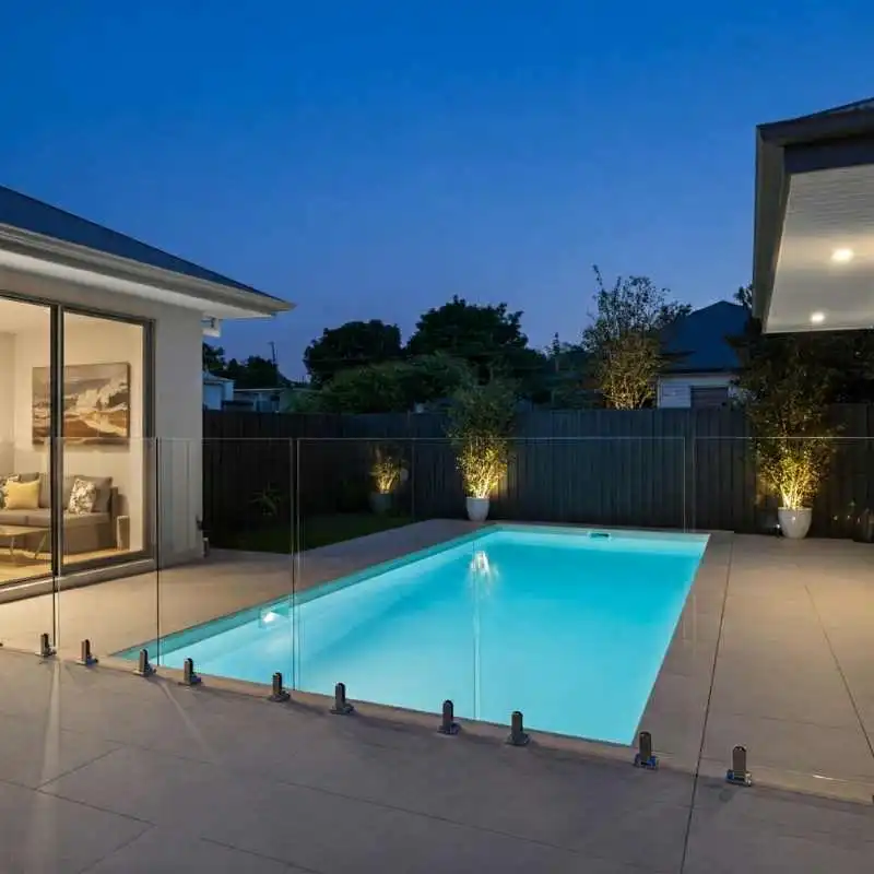 A rectangular magnesium swimming pool glows with soft blue light at dusk. The pool is enclosed by a frameless glass fence and set within a large tiled patio. In the background, a dark privacy fence is lined with small trees illuminated by warm garden uplighting.