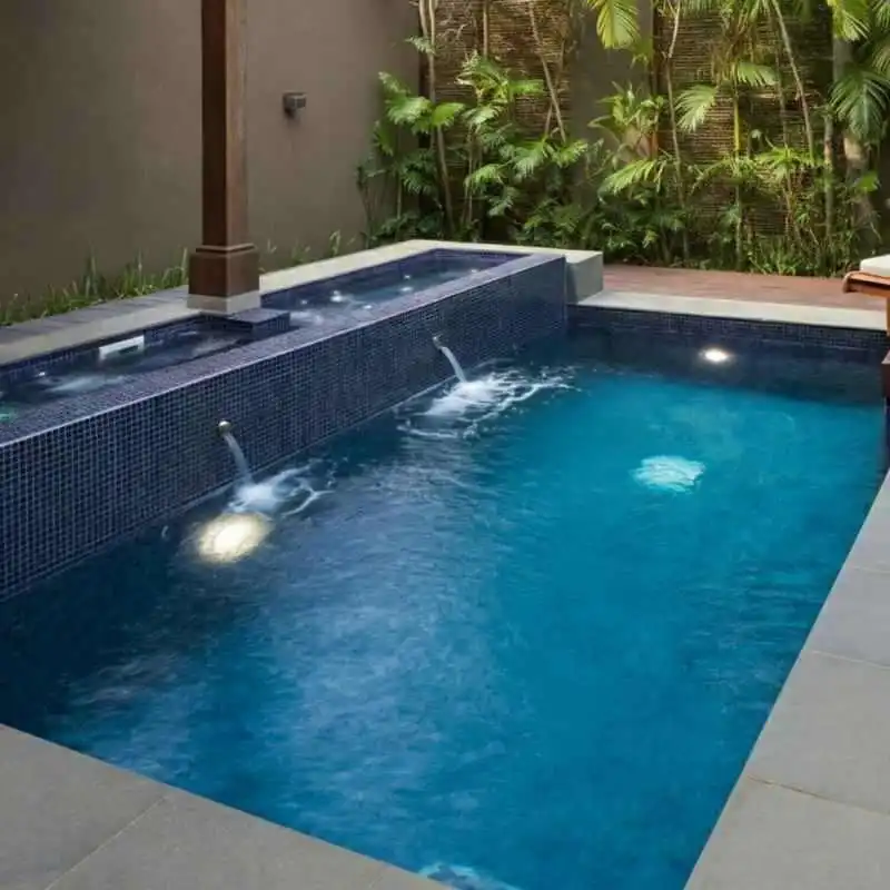 A narrow magnesium lap pool features deep blue mosaic tiling and two stainless steel water spouts pouring into the water. The pool is illuminated by underwater lights and set against a neutral-toned wall with lush tropical palms and a wooden deck in the background.