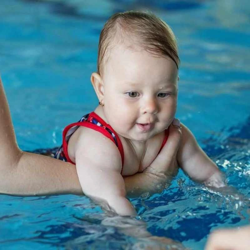 A close-up shot of a baby being supported by an adult’s hands while splashing in the clear blue water of a swimming pool. The baby is wearing a red and blue patterned swimsuit and looking down at the water.